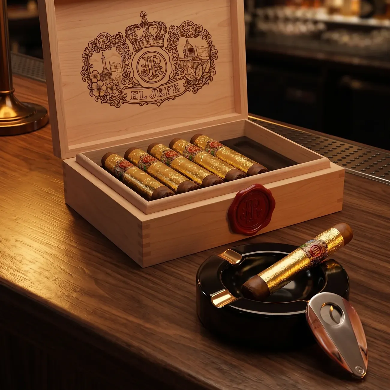 An open light-wood El Jefe box of gold-foil cigars with red bands on a bar counter, with another gold-foil cigar on a black ashtray and a cigar cutter in front, a table lamp and bar shelves behind