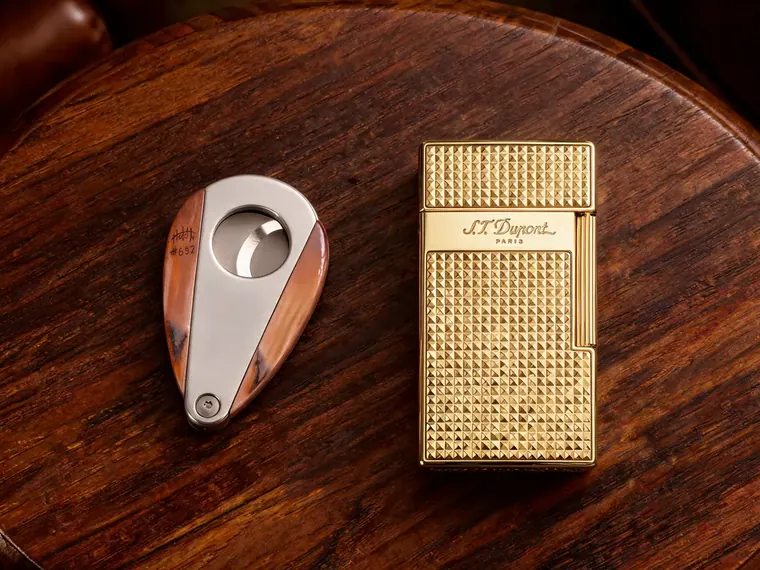 A steel and wood cigar cutter beside a gold diamond-pattern lighter on a round wooden table