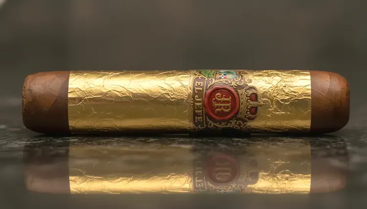 A gold-leaf wrapped El Jefe cigar with the red wax-seal JR band on a dark reflective surface